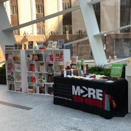 A table covered with a black tablecloth with the More Than Words logo sits in front of a white bookcase.