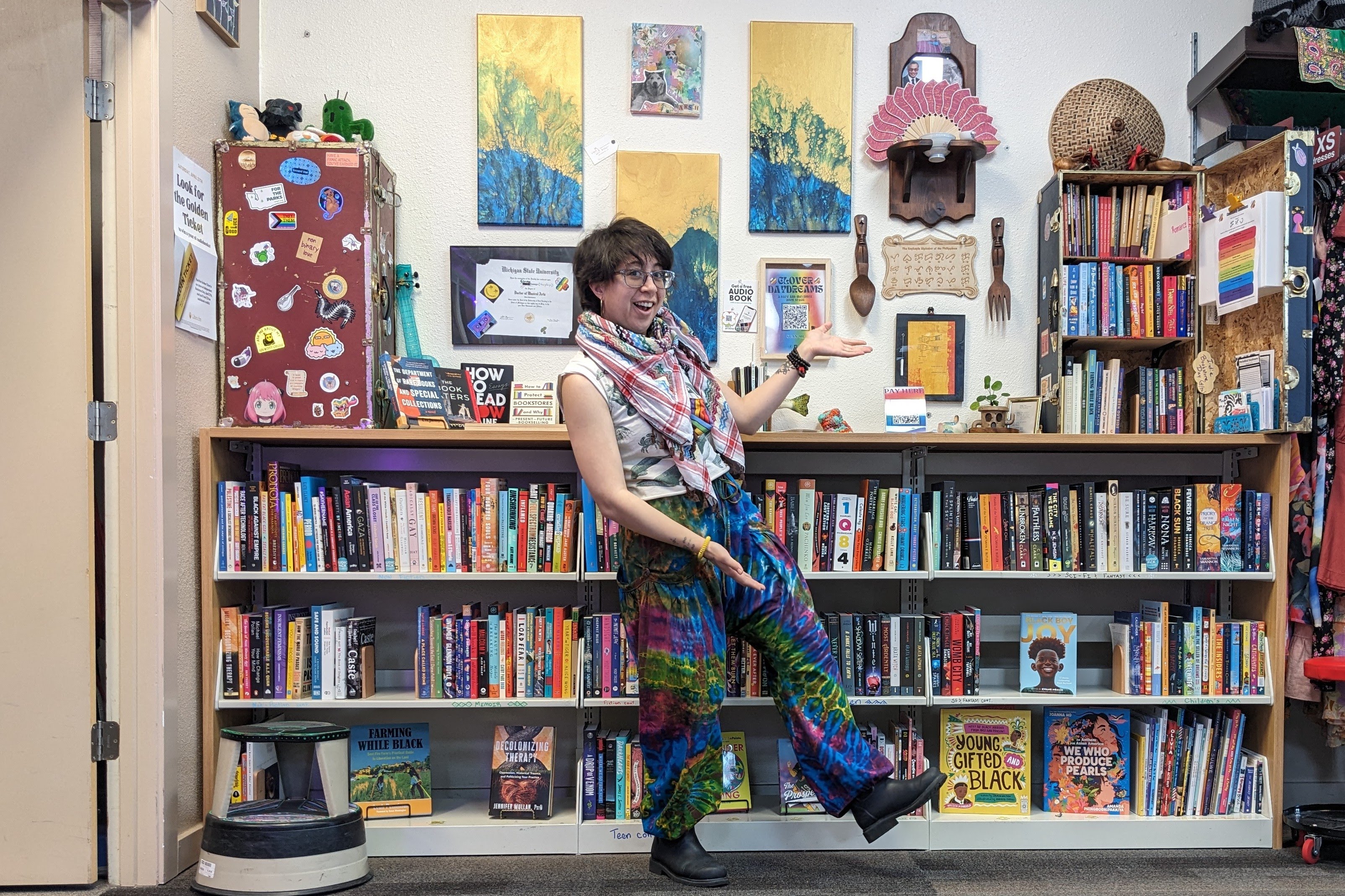 Clover of Clover Daydreams stands in front of a bookshelf and gestures to one of the steamer trunks that has been converted into a bookcase.