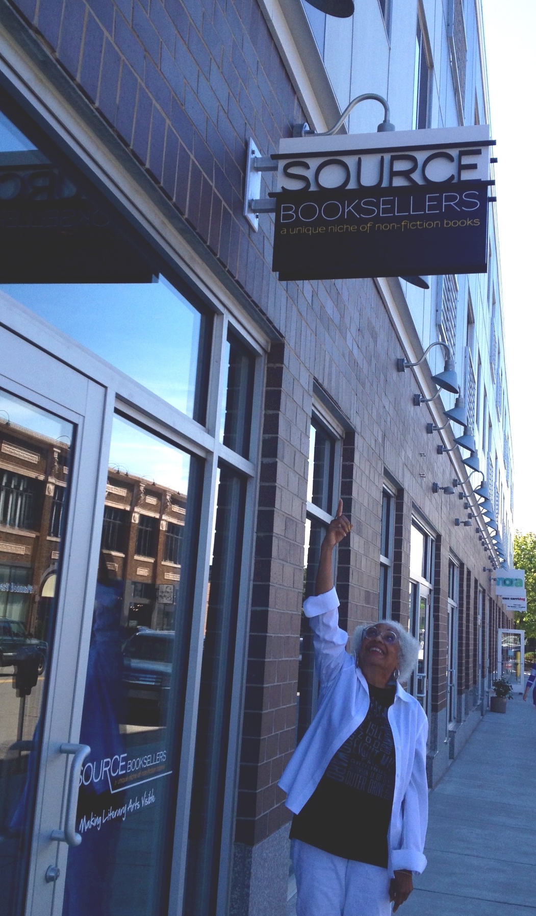 Janet Webster Jones stands outside Source Booksellers, pointing up at the store's sign.