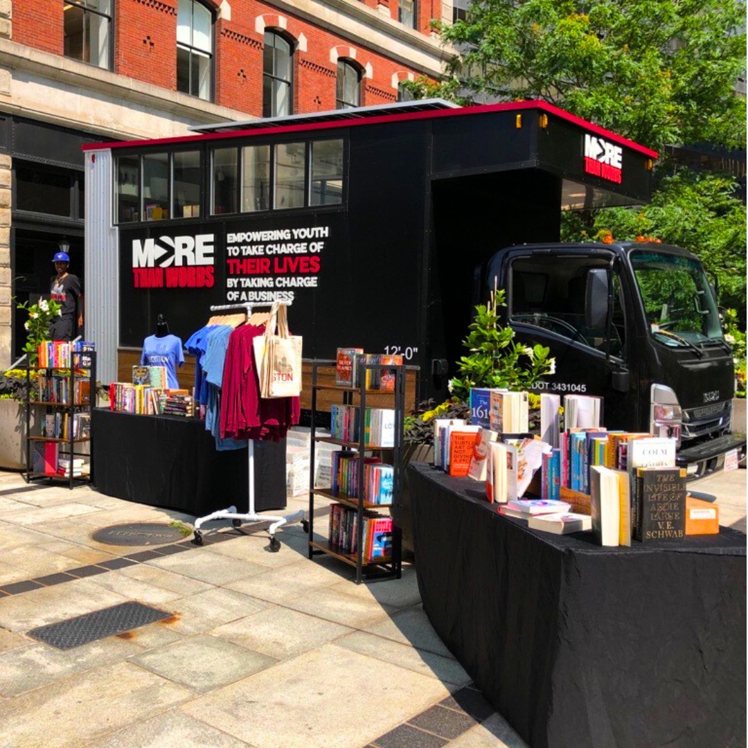 A large book bus with the More Than Words logo sits behind shelves of books and racks of clothing.