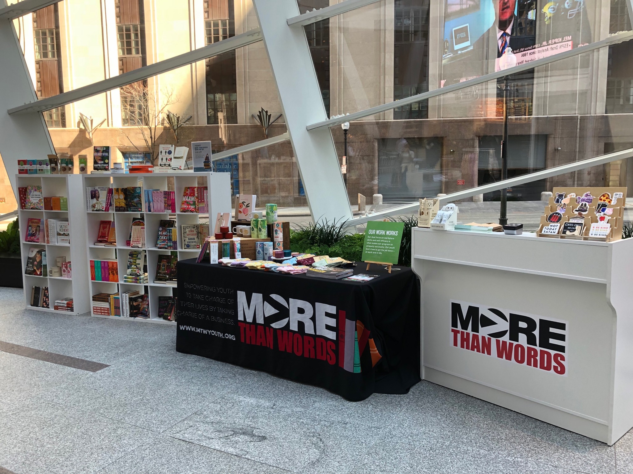 A table with a More Than Words table cloth is covered in books. It sits next to a full bookshelf.