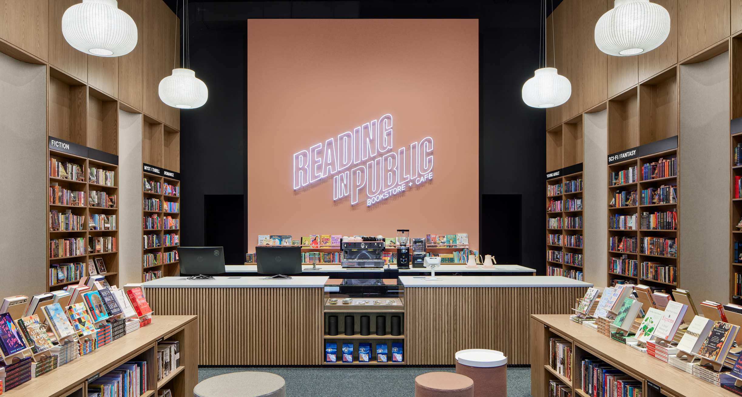 Rows of low bookshelves lead to a counter with cash registers in front of a pale pink wall. A white neon sign in the wall says "Reading in Public."